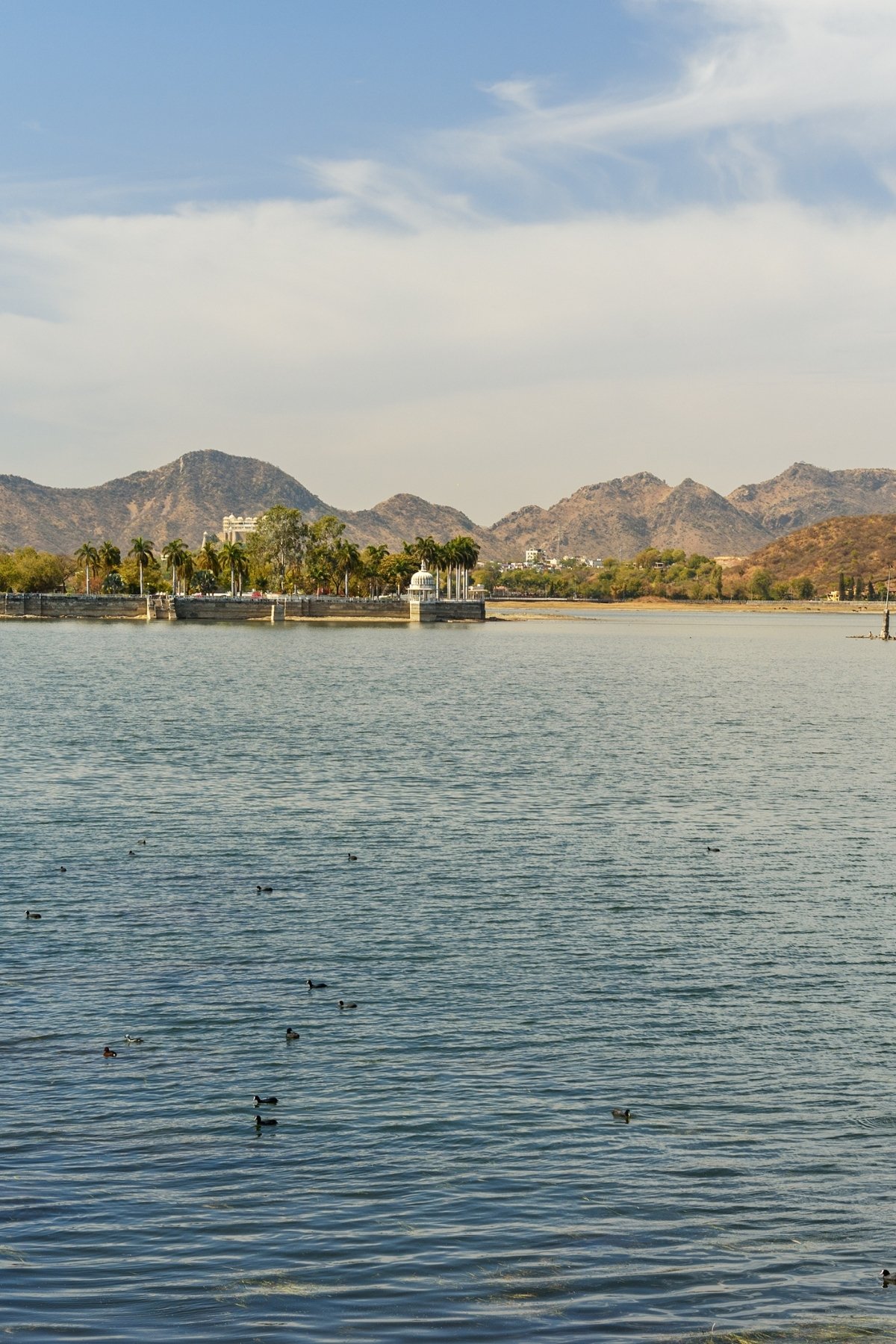 Fateh Sagar Lake – Where Water Holds the Sky and Silence Finds Shape. Fateh Sagar Lake in Udaipur, a tranquil blue expanse surrounded by Aravalli hills, with motorboats gliding across and Nehru Park Island nestled at its heart.